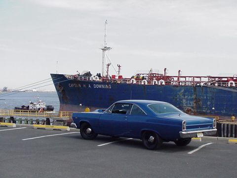 1966 Ford Fairlane - A blue 1966 Ford Fairlane coupe is parked in front of a large cargo ship at a dock.