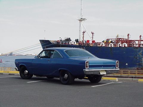 1966 Ford Fairlane - A blue 1966 Ford Fairlane coupe is parked near a large cargo ship at a waterfront.