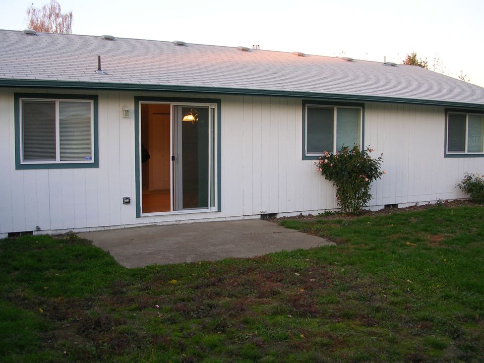 White house exterior with a sliding glass door and two windows, a concrete patio, and grass