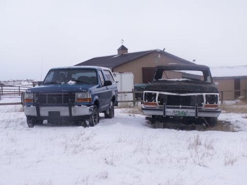 1995 Ford Bronco - Two Ford trucks, one blue Bronco and one damaged dark truck, sit in a snowy landscape in front of a barn.