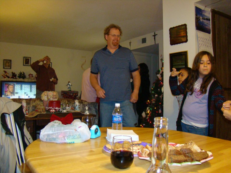 Interior scene with people gathered around a table with food and drinks, a Christmas tree in the background