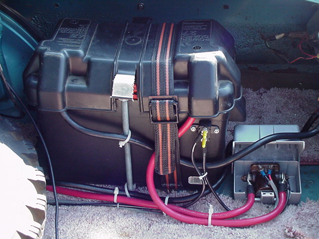 Dual battery box with red and black cables and relay mounted in vehicle cargo area on carpet