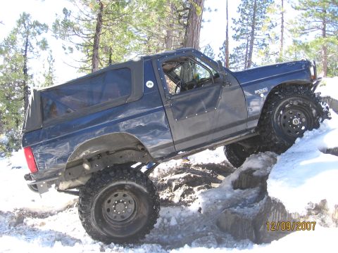 1994 Ford Bronco - A blue Ford Bronco navigates a snowy, rocky trail. The vehicle's badge indicates it is a Bronco.