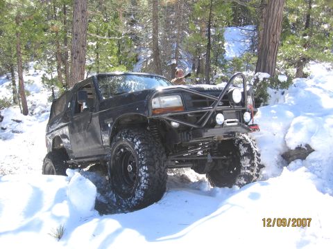 1994 Ford Bronco - Modified black Ford Bronco navigates a snowy, wooded terrain with a custom roll cage and auxiliary lights.