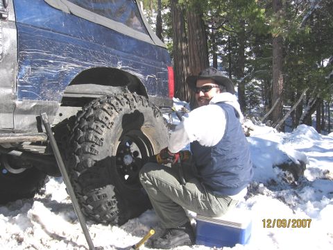 1994 Ford Bronco - A person works on a flat tire of a blue SUV in a snowy, wooded area.