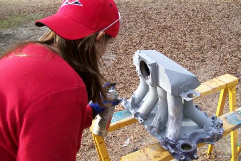 1988 Ford Bronco - A person in a red cap and shirt spray paints a gray intake manifold, which is resting on sawhorses outdoors.