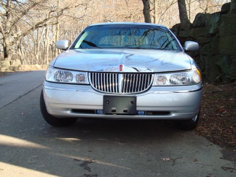 2002 Lincoln Town Car - A silver Lincoln Town Car is parked on a road with a stone wall and trees in the background.