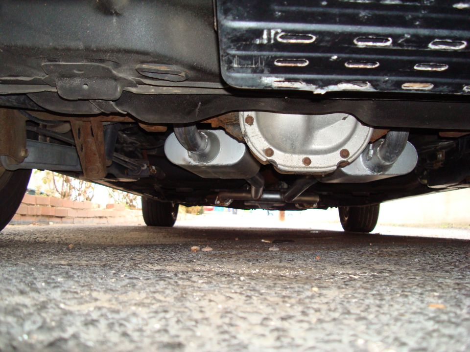 Underside view of car's rear differential and dual mufflers with exhaust piping over wet asphalt