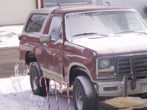 1986 Ford Bronco - A maroon Ford Bronco sits covered in snow, with holiday decorations nearby.