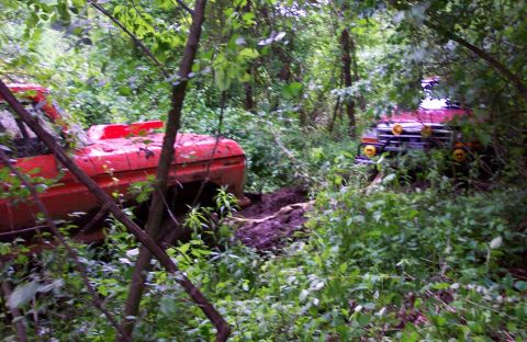 1995 Ford Bronco - Two off-road trucks are stuck in the mud, surrounded by dense foliage and trees.