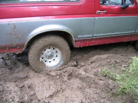 1995 Ford Bronco - This red and silver SUV, likely a 1992 model, is deeply embedded in thick mud, with only its side profile and front wheel visible.