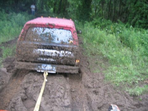 1995 Ford Bronco - A red SUV is deeply stuck in thick mud, with a tow strap attached to its rear bumper.
