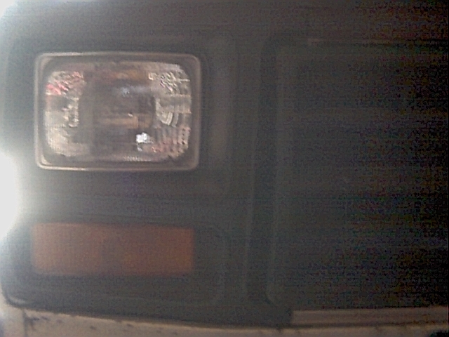 Dark, blurry close-up of a square sealed-beam headlight and amber marker on a vehicle