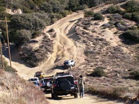 2007 Toyota FJ Cruiser - Several SUVs, including a black 2007 Toyota FJ Cruiser, navigate a steep, winding dirt path in a hilly, arid landscape.