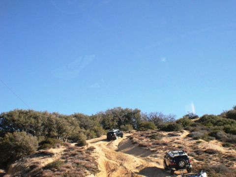 2007 Toyota FJ Cruiser - Two white SUVs navigate a challenging off-road trail, kicking up dust under a bright blue sky.