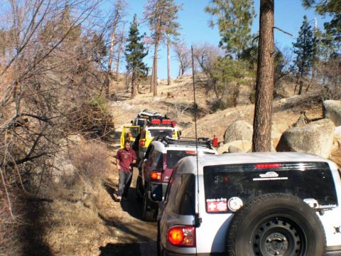 2007 Toyota FJ Cruiser - Several Toyota FJ Cruisers are seen on an off-road trail, navigating a rocky and wooded terrain.