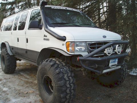 2000 Ford E350 - A white lifted Ford E-Series van photographed from the front three-quarter angle, riding on oversized off-road tires with a black tube fron…