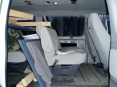 2000 Ford E350 - The interior of a van shows passenger seats and makeshift wooden shelving, with camouflage fabric visible.