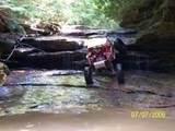 1992 Ford Mustang - An off-road vehicle makes its way through a shallow stream surrounded by rocks and foliage.