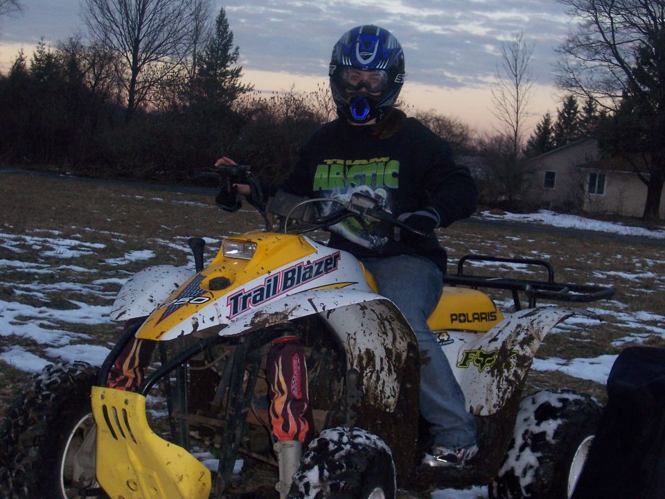 Rider in helmet on muddy yellow Polaris Trail Blazer ATV in winter field