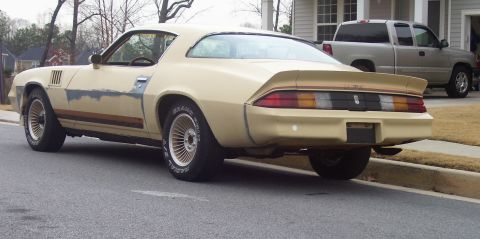 1979 Chevrolet Camaro - A tan 1979 Chevrolet Camaro with visible body damage and racing stripes is parked on the street, with a pickup truck in the background.