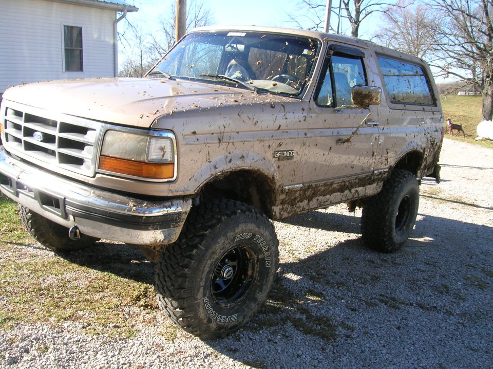 Tan Ford Bronco covered in mud, lifted with off-road tires, parked on gravel