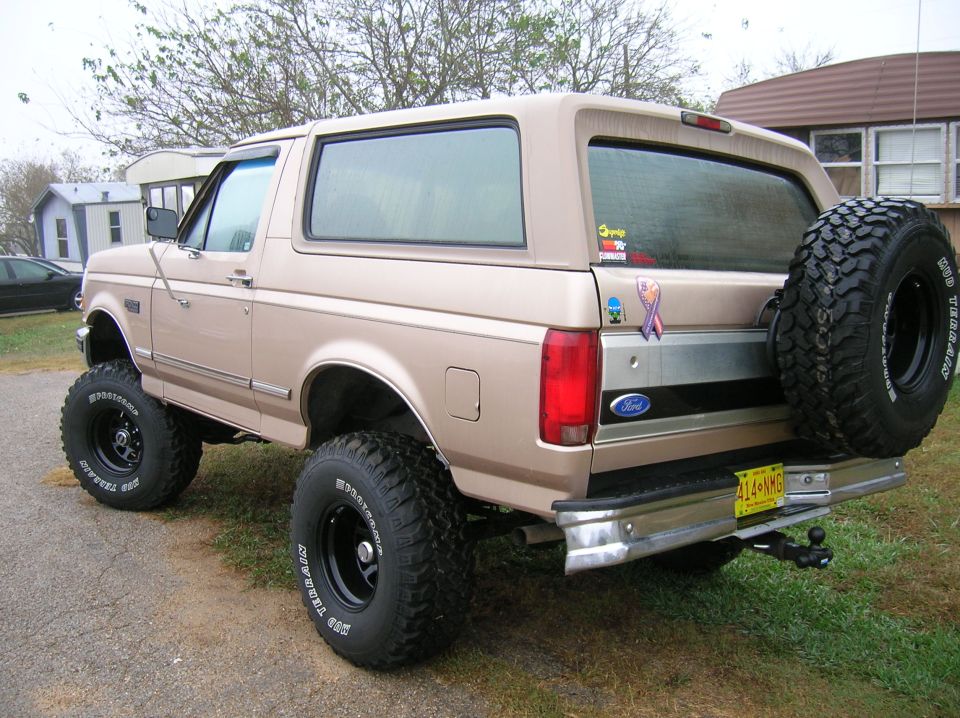 Tan Ford Bronco, lifted with big tires, spare tire on back. Yellow license plate visible