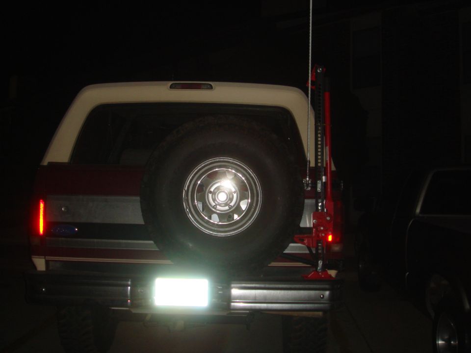 Rear of maroon and tan Ford Bronco at night with spare tire and red Hi-Lift jack mounted on tailgate