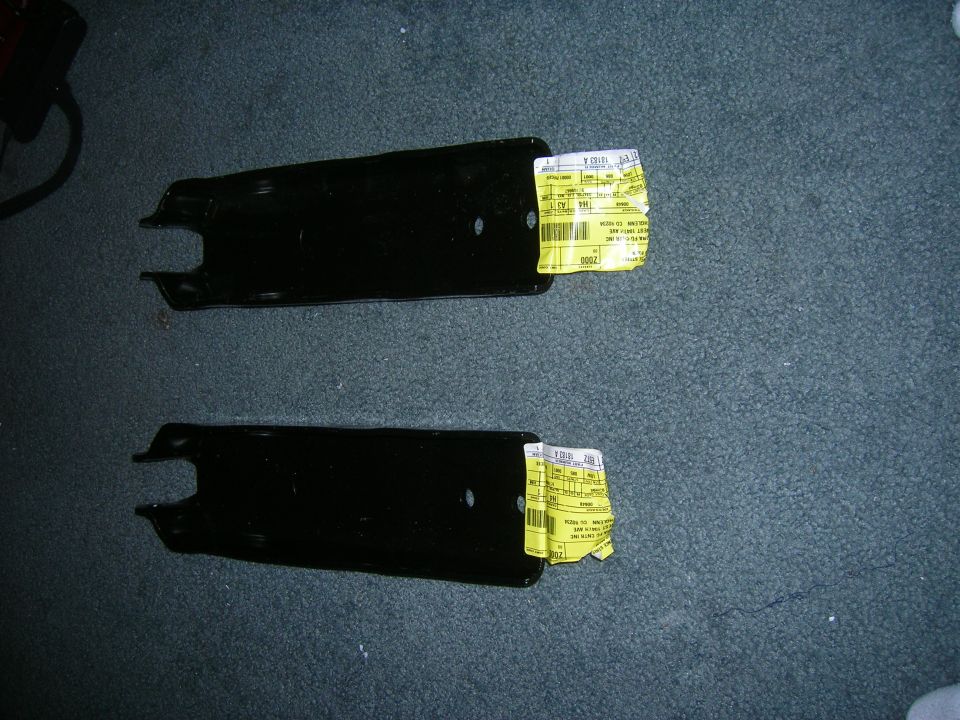 Two black metal suspension components with yellow labels attached, lying on a carpeted surface