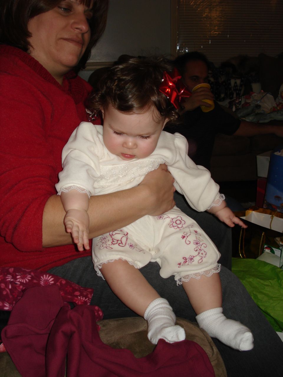 Woman holding a baby with a red bow in her hair. Another person is in the background
