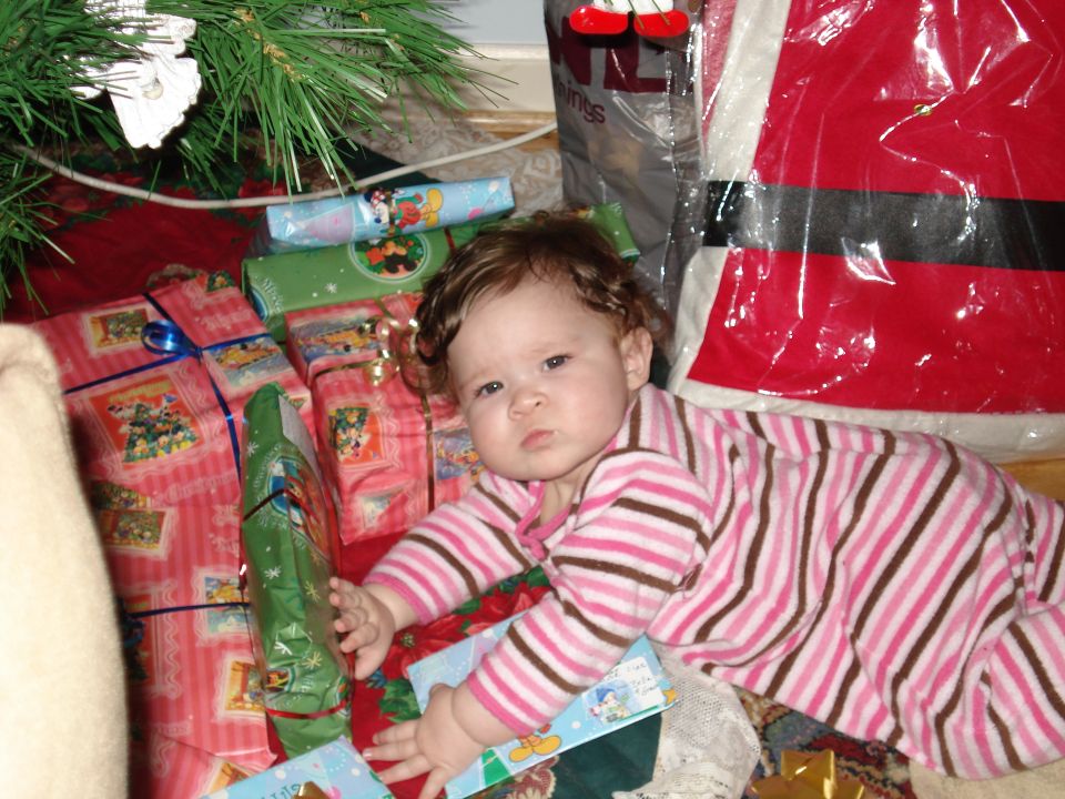 Baby in pink striped pajamas lies on the floor near a Christmas tree and wrapped gifts