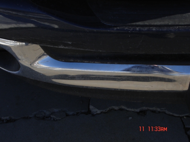 Close-up of a chrome bumper with an exhaust pipe visible on the left. The reflection on the bumper shows a landscape