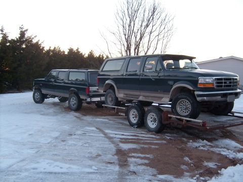 1995 Ford C-350 (Centurion Conversions) - A Ford Bronco SUV is loaded onto a trailer, with a Ford F-Series truck following behind it in a snowy outdoor setting.