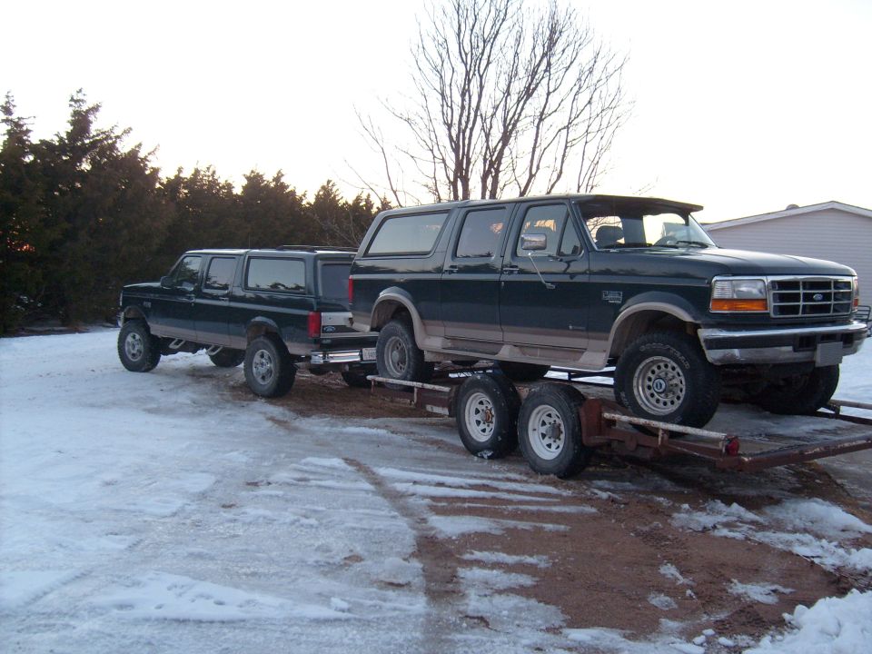 Green and tan Ford Bronco on tandem trailer behind a green crew cab Ford pickup in snow