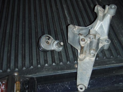 1995 Ford F-150 - A belt tensioner and its mounting bracket are shown resting on a dark, ribbed surface, possibly a truck bed.