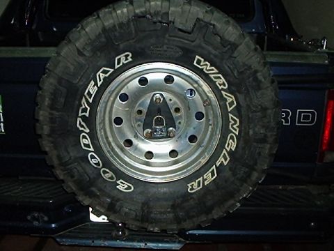 1995 Ford F-150 - A Goodyear Wrangler mud-terrain tire with raised white lettering on a polished ten-hole aluminum wheel, mounted as a spare against the tail…
