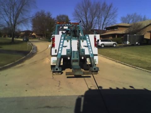 1992 Ford Bronco - A tow truck is seen from the rear on a residential street, with another vehicle and a trailer visible in the background.