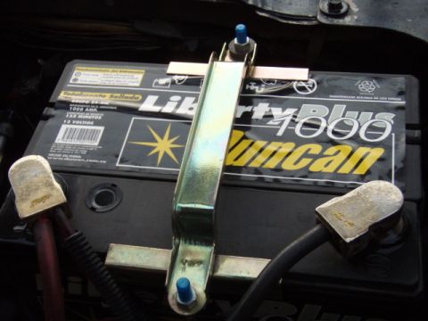1995 Ford F-150 - Close-up of a car battery and its hold-down bracket, with visible text on the battery label.