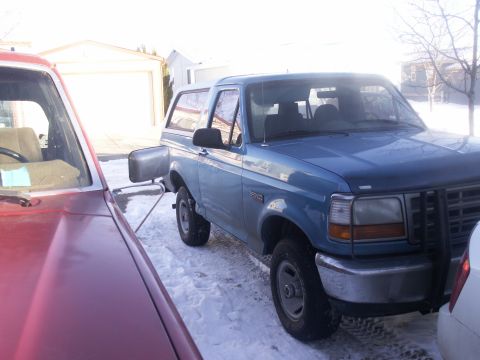 1995 Ford Bronco - A blue Ford Bronco is parked in the snow, partially obscured by a red vehicle.