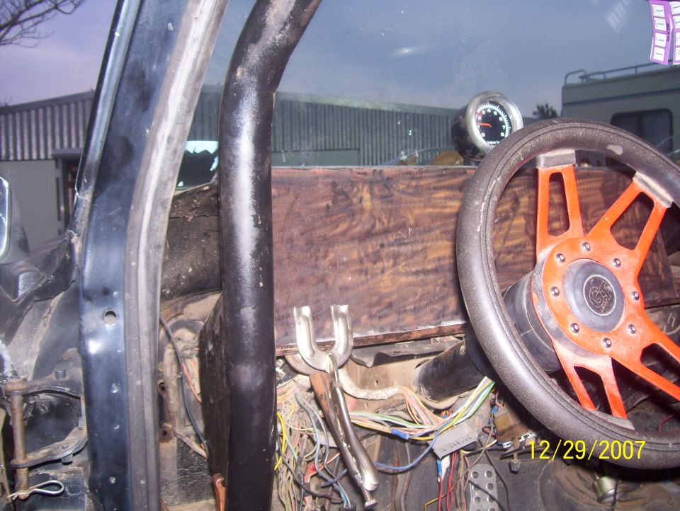 Interior view of a vehicle with a custom dashboard, aftermarket orange steering wheel, and a gauge. Wires are visible