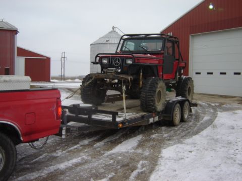 1992 Ford Bronco - A red off-road vehicle is loaded onto a trailer, hitched to a red pickup truck, with farm buildings in the background.