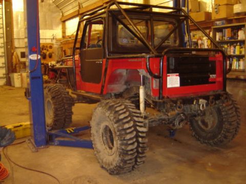 1992 Ford Bronco - Modified red off-road vehicle with a prominent roll cage and oversized tires is shown on a hydraulic lift in a garage setting.