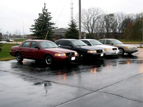 2001 Ford Crown Victoria - A line of four Ford Crown Victorias, including a red one in the foreground, are parked on a wet asphalt lot under an overcast sky.