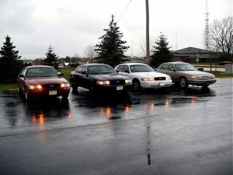 2001 Ford Crown Victoria - Four Ford Crown Victorias are parked in a lot on a cloudy, rainy day, with their headlights on.
