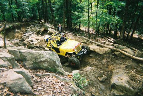 1976 Ford Bronco - A yellow off-road vehicle navigates a challenging rocky path in a wooded area.