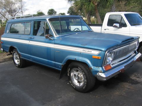 1978 Jeep Cherokee - This blue Jeep Wagoneer features a distinctive chrome grille and side trim, with a white pickup truck visible in the background.