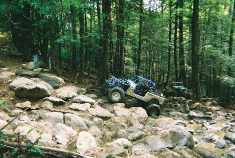 1976 Ford Bronco - Off-road vehicles tackle a challenging rocky path through a dense forest.