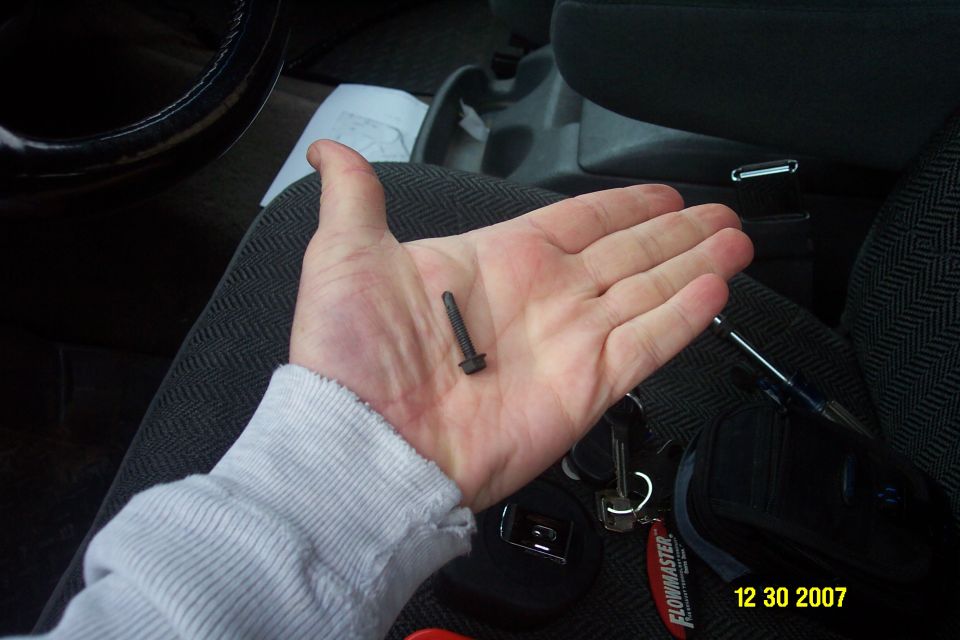 Hand holding a small black flange bolt inside a vehicle, keys and tools on gray seat