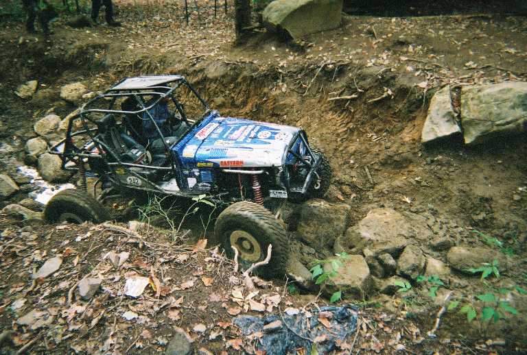 Off-road buggy with a roll cage navigating a rocky, muddy ditch. It has blue and white body panels with some text and logos