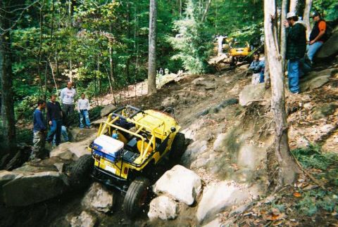 1976 Ford Bronco - A group of people watch a yellow off-road vehicle tackle a challenging rocky trail in a wooded area.